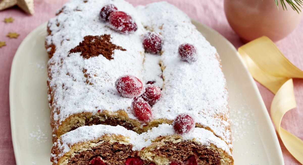 Festlicher Spekulatius-Stollen mit Puderzucker und kandierten Cranberrys auf Platte, daneben goldene Deko auf rosa Stofftischdecke.