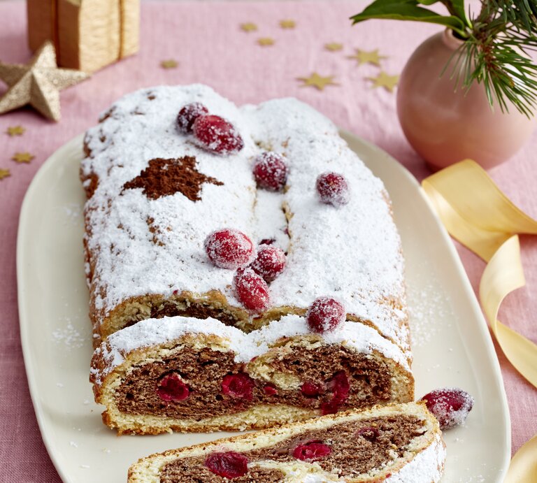 Festlicher Spekulatius-Stollen mit Puderzucker und kandierten Cranberrys auf Platte, daneben goldene Deko auf rosa Stofftischdecke. | © EAT CLUB / Isabell Triemer