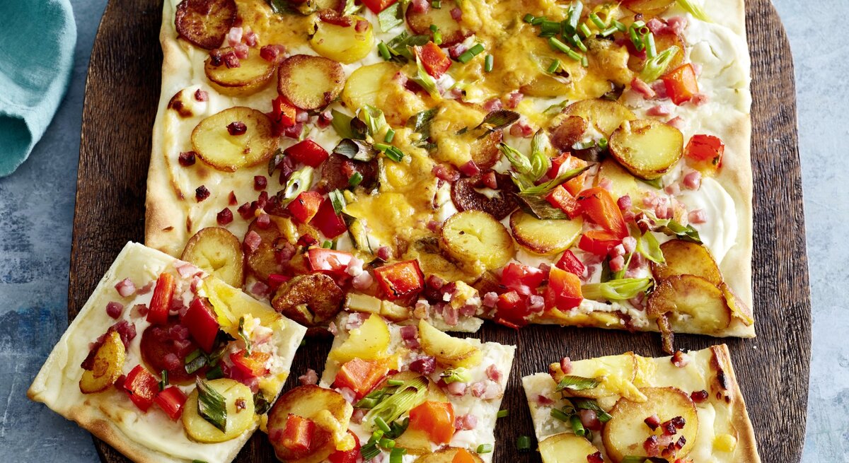 Speck-Flammkuchen auf einem hölzernem Blech auf grauem Hintergrund. 