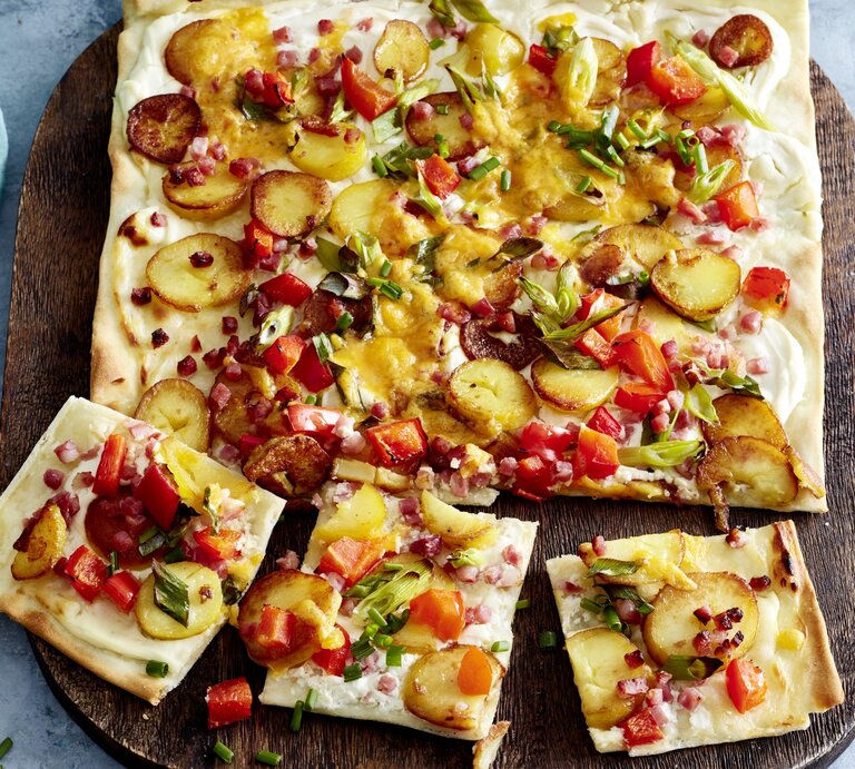 Speck-Flammkuchen auf einem hölzernem Blech auf grauem Hintergrund.  | © EAT CLUB/Isabell Triemer