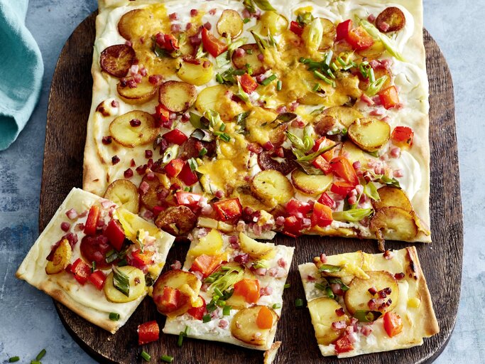 Speck-Flammkuchen auf einem hölzernem Blech auf grauem Hintergrund.  | © EAT CLUB/Isabell Triemer