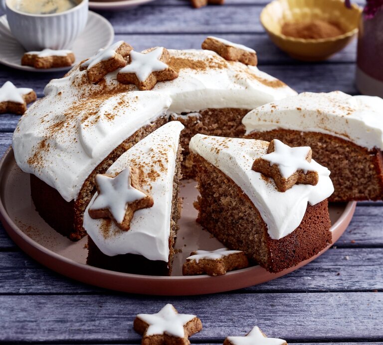 Zimtstern-Nusskuchen auf einem braunen Teller auf grauem Hintergrund. Links hinten auf dem Foto eine Tasse Kaffee. | © EAT CLUB / Isabell Triemer
