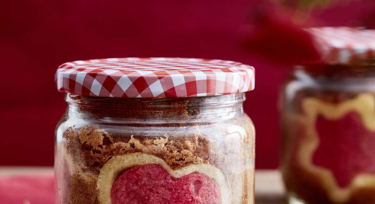 Selbstgebackener Kuchen mit rotem Herz in einem Schraubglas mit Karodeckel, daneben ein Geschenkanhänger auf Holz.