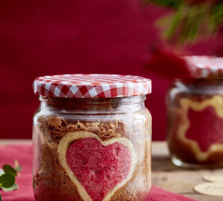 Selbstgebackener Kuchen mit rotem Herz in einem Schraubglas mit Karodeckel, daneben ein Geschenkanhänger auf Holz. | © EAT CLUB/Isabell Triemer