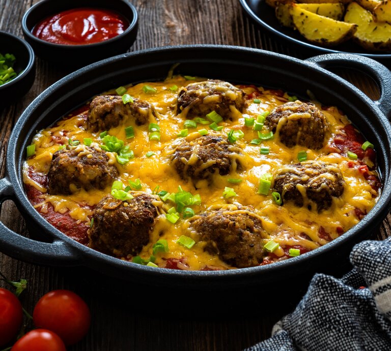 Überbackene Cowboy Fleischbällchen mit geschmolzenem Käse und grünen Zwiebeln in Tomatensauce, serviert auf rustikalem Holztisch. | © Shutterstock/Jacek Chabraszewski