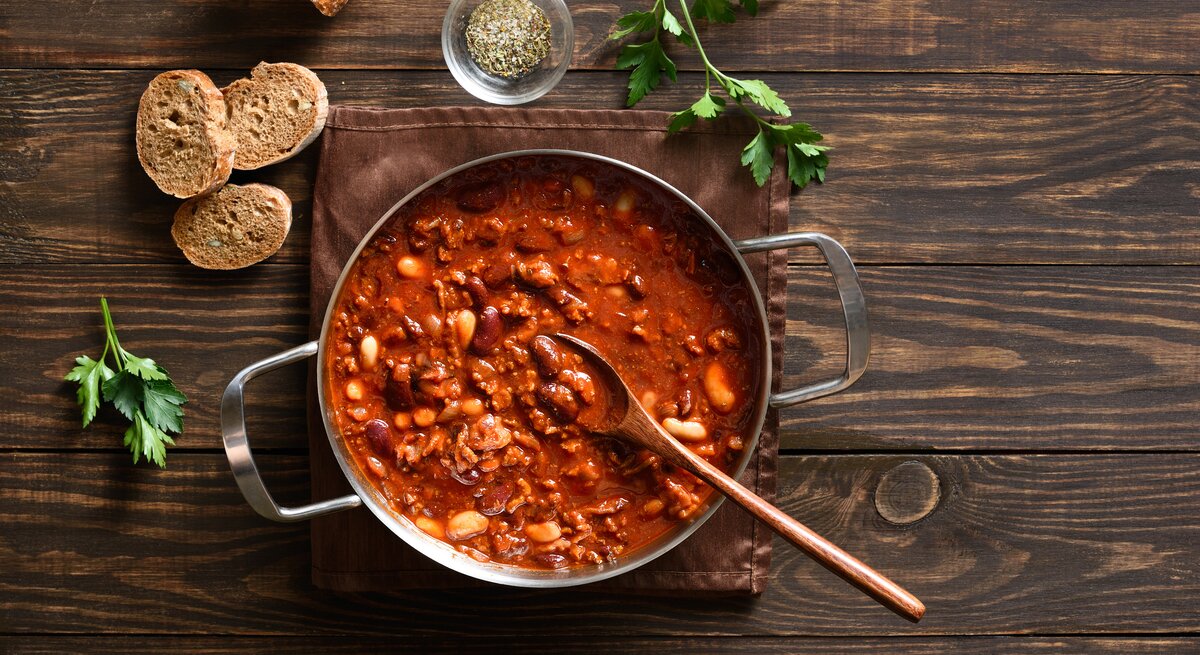 Deftige Cowboy Bohnen mit Hackfleisch und Bohnenmix in roter Sauce, angerichtet im Topf auf rustikalem Holztisch.