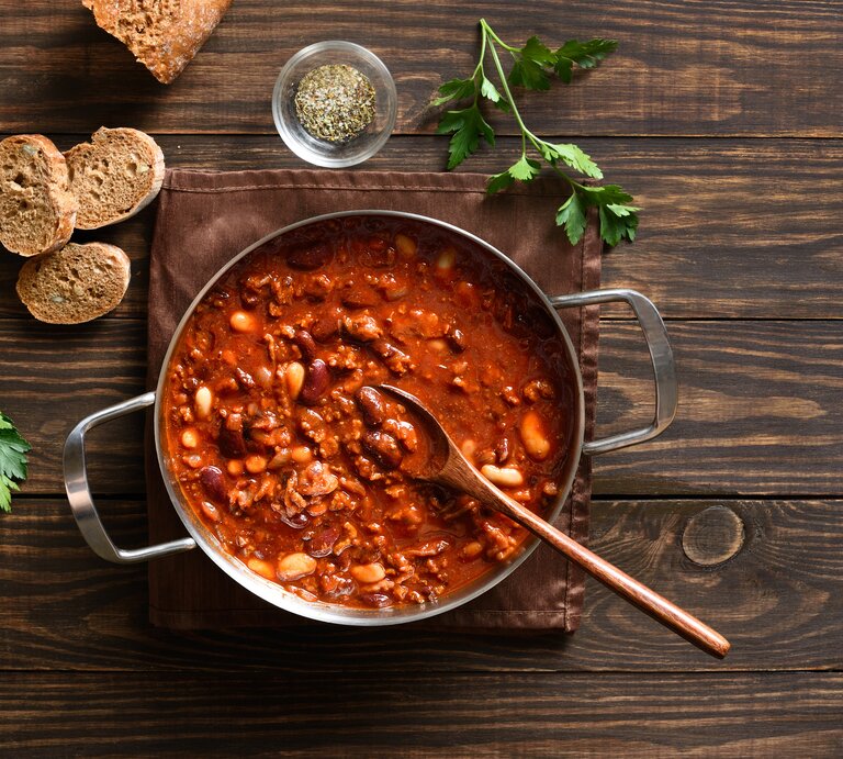 Deftige Cowboy Bohnen mit Hackfleisch und Bohnenmix in roter Sauce, angerichtet im Topf auf rustikalem Holztisch. | © Shutterstock/Tatiana Volgutova