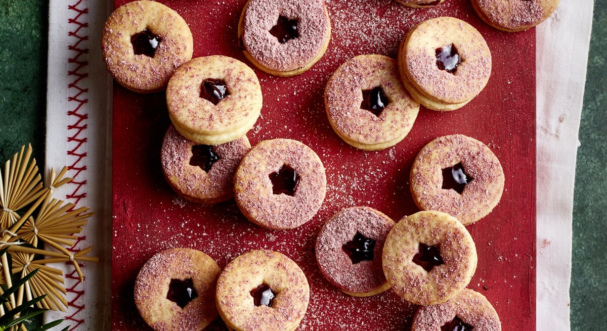 Brombeer-Marzipan-Kekse mit dunkler Marmelade und rosafarbenem Zucker auf rustikalem Brett, daneben Sieb und Deko.