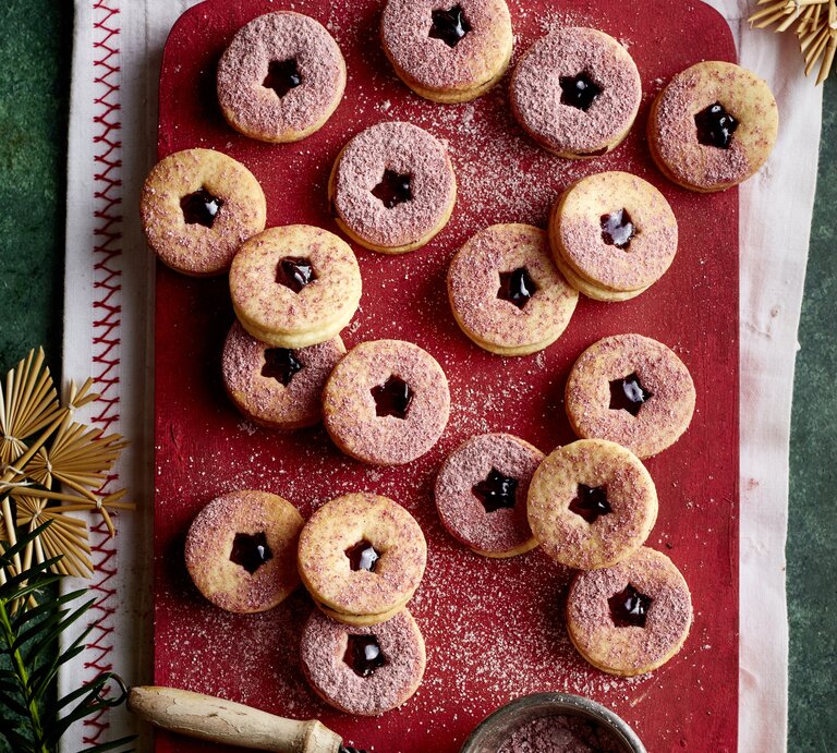 Brombeer-Marzipan-Kekse mit dunkler Marmelade und rosafarbenem Zucker auf rustikalem Brett, daneben Sieb und Deko. | © EAT CLUB/Isabell Triemer