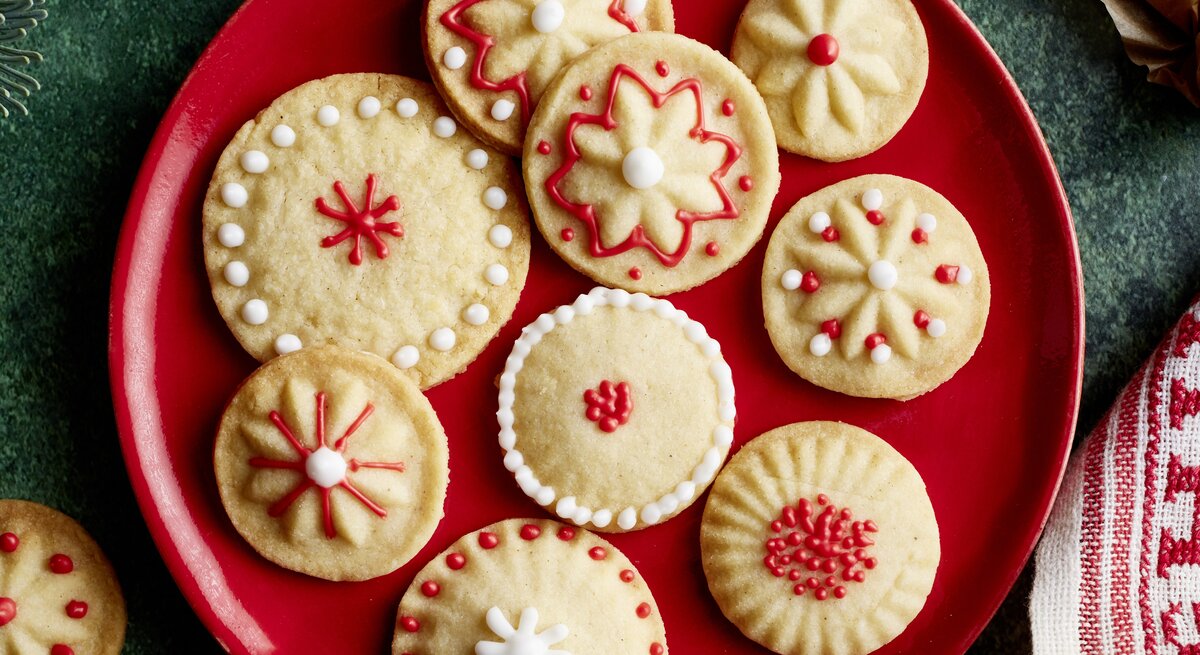 Elf liebevoll verzierte Vanille-Plätzchen mit filigranen Mustern in Rot und Weiß auf rotem Teller auf grünem Untergrund.