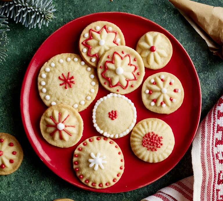 Elf liebevoll verzierte Vanille-Plätzchen mit filigranen Mustern in Rot und Weiß auf rotem Teller auf grünem Untergrund. | © EAT CLUB/Isabell Triemer