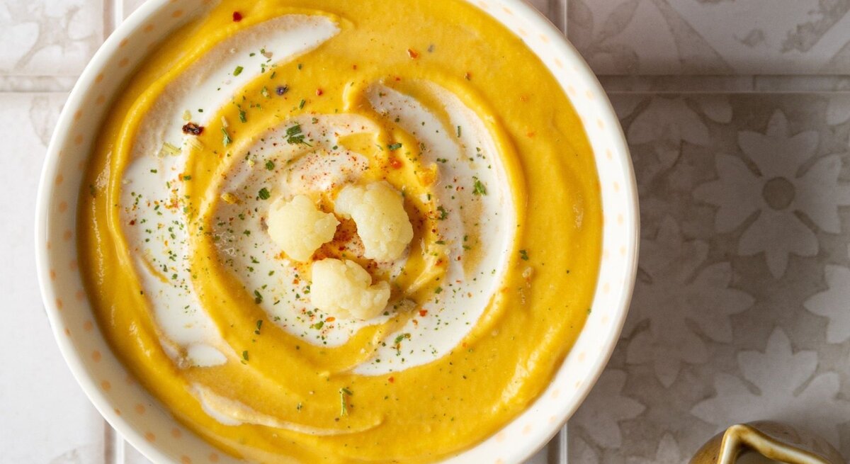 Zwei Schüsseln mit cremiger Kürbis-Blumenkohl-Suppe, dekoriert mit einem Swirl aus Joghurt und gerösteten Blumenkohlröschen, auf einer hellen, gemusterten Fliesenoberfläche. Neben den Schüsseln steht eine mit Suppe gefüllte Pfanne und eine kleine Kanne, umgeben von beigen Stoffservietten.