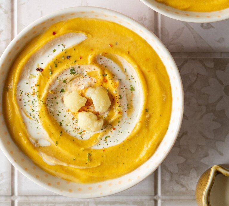 Zwei Schüsseln mit cremiger Kürbis-Blumenkohl-Suppe, dekoriert mit einem Swirl aus Joghurt und gerösteten Blumenkohlröschen, auf einer hellen, gemusterten Fliesenoberfläche. Neben den Schüsseln steht eine mit Suppe gefüllte Pfanne und eine kleine Kanne, umgeben von beigen Stoffservietten. | © Adobe Stock / lacemika