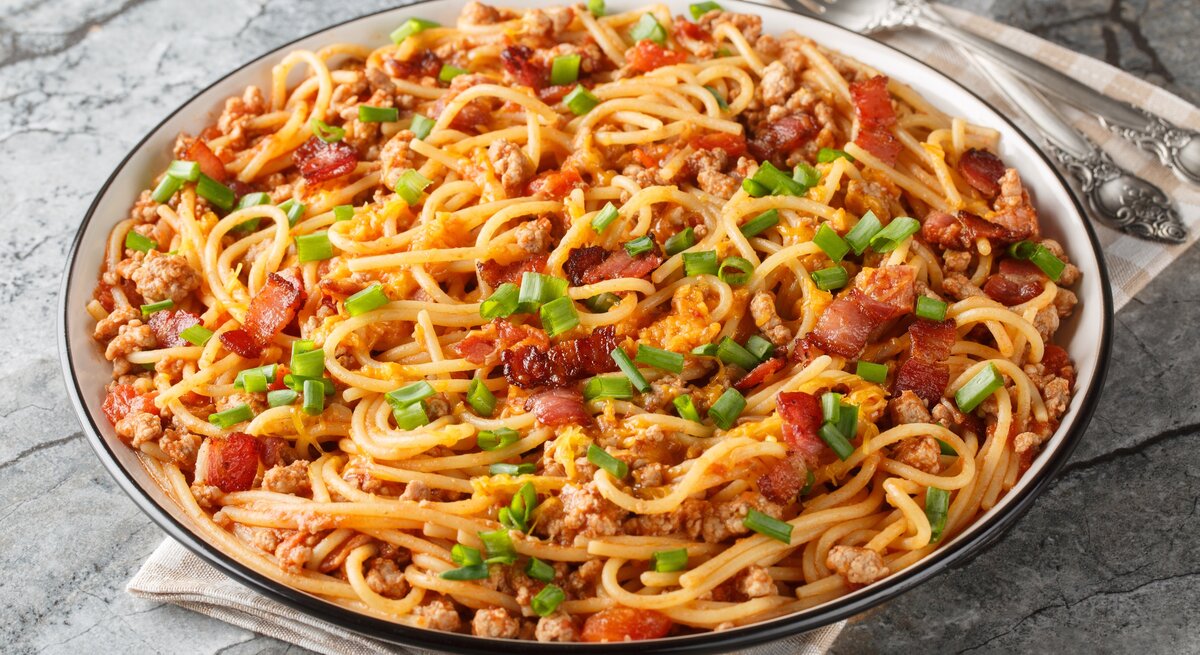 Cowboy Spaghetti mit Hackfleisch, Speck, Tomatenstücken und Frühlingszwiebeln auf einem Teller mit Besteck auf Marmorplatte.