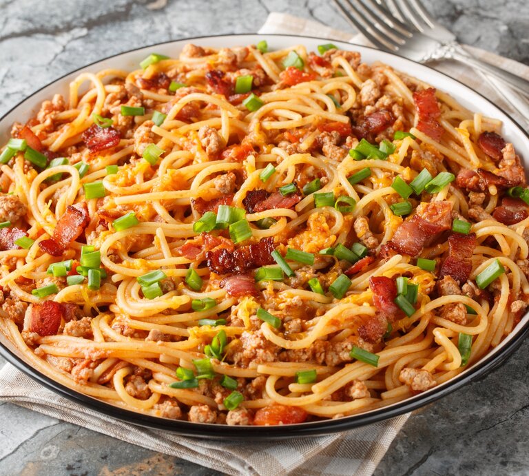 Cowboy Spaghetti mit Hackfleisch, Speck, Tomatenstücken und Frühlingszwiebeln auf einem Teller mit Besteck auf Marmorplatte. | © Shutterstock/Sergii Koval