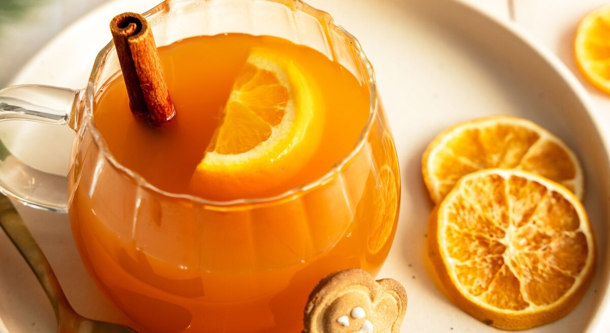 Ein Glas mit alkoholfreier Orangenpunsch, dekoriert mit einer Orangenscheibe und einer Zimtstange, steht auf einem Teller. Neben dem Glas liegen getrocknete Orangenscheiben, ein goldener Löffel und ein Lebkuchenplätzchen in Form eines Lebkuchenmanns. Die warme und gemütliche Szene vermittelt Winter- und Weihnachtsstimmung.