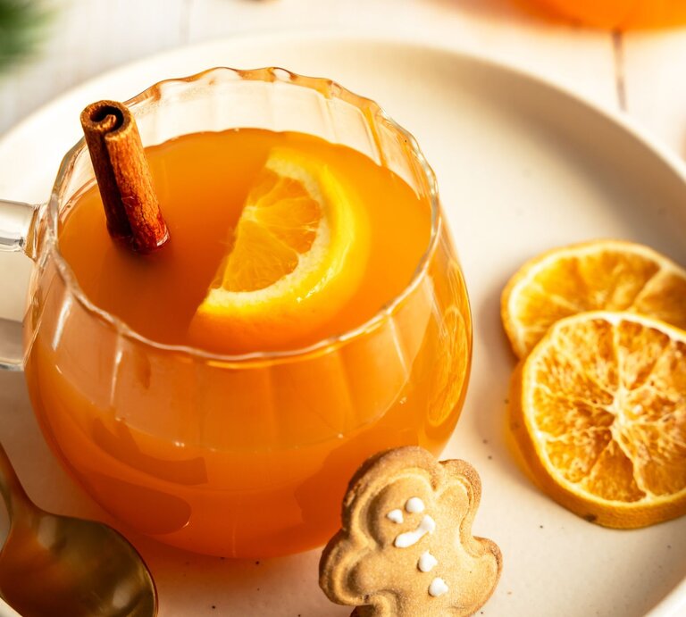 Ein Glas mit alkoholfreier Orangenpunsch, dekoriert mit einer Orangenscheibe und einer Zimtstange, steht auf einem Teller. Neben dem Glas liegen getrocknete Orangenscheiben, ein goldener Löffel und ein Lebkuchenplätzchen in Form eines Lebkuchenmanns. Die warme und gemütliche Szene vermittelt Winter- und Weihnachtsstimmung. | © Adobe Stock / Sonja Rachbauer