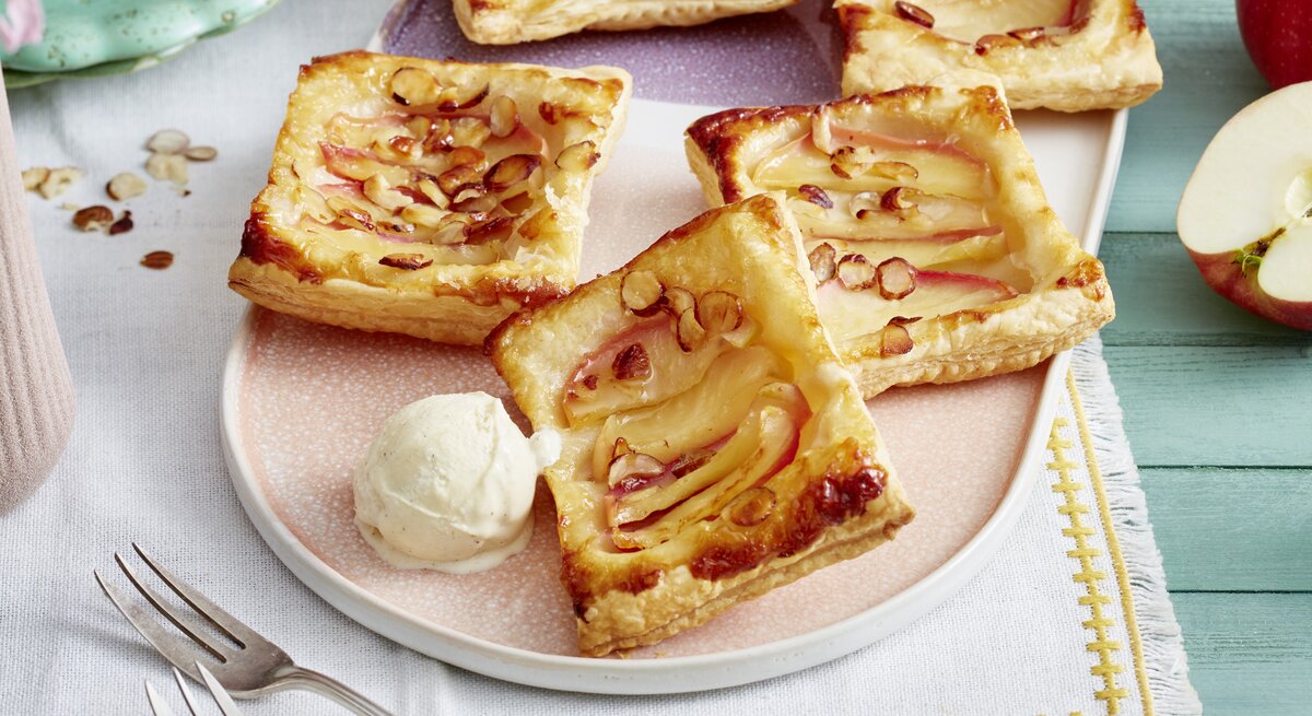 Vier Blätterteigtörtchen mit Apfelscheiben und gehobelten Mandeln liegen auf einem Teller neben einer Kugel Vanilleeis.