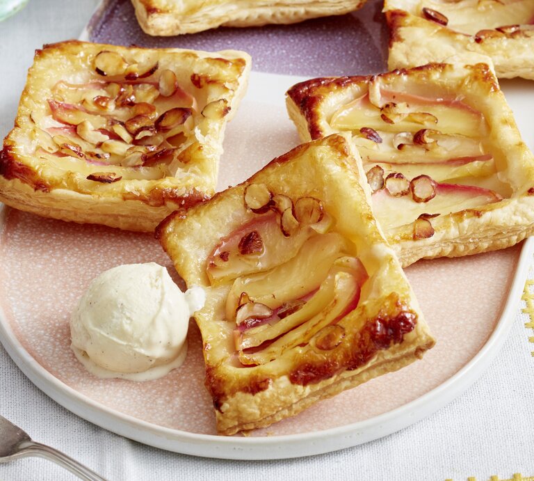 Vier Blätterteigtörtchen mit Apfelscheiben und gehobelten Mandeln liegen auf einem Teller neben einer Kugel Vanilleeis. | © EAT CLUB/Isabell Triemer
