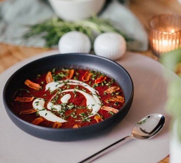 Eine Schale Rotkohl-Schmand-Suppe mit Karotten-Chips mit dekorativem Sahne-Wirbel, gehackten Kräutern und knusprigen Topping-Streifen, serviert auf einem gedeckten Tisch mit Kerzenlicht und einem Löffel daneben. | © Rabenhorst