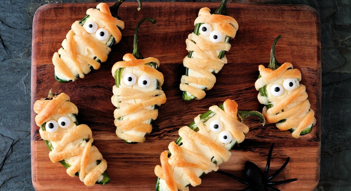Sechs Mumien Jalapeno Popper auf einem Holzbrett – halbierte Jalapeños, mit Frischkäse gefüllt, in Teigstreifen gewickelt und mit Zuckeraugen dekoriert, als lustiger Halloween-Snack angerichtet.