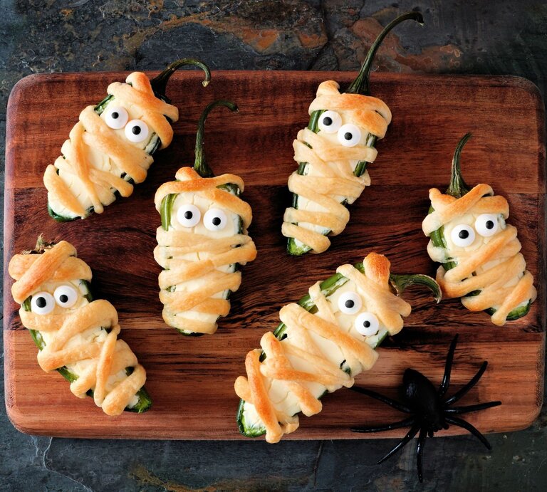 Sechs Mumien Jalapeno Popper auf einem Holzbrett – halbierte Jalapeños, mit Frischkäse gefüllt, in Teigstreifen gewickelt und mit Zuckeraugen dekoriert, als lustiger Halloween-Snack angerichtet. | © Uhr AdobeStock/Jenifoto