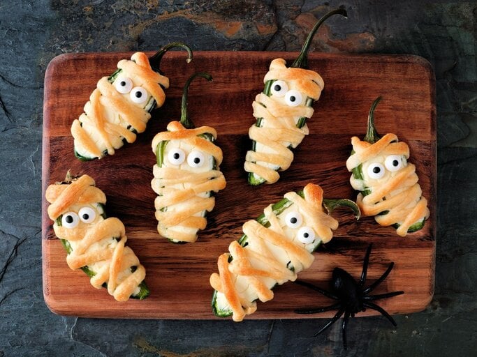 Sechs Mumien Jalapeno Popper auf einem Holzbrett – halbierte Jalapeños, mit Frischkäse gefüllt, in Teigstreifen gewickelt und mit Zuckeraugen dekoriert, als lustiger Halloween-Snack angerichtet. | © Uhr AdobeStock/Jenifoto