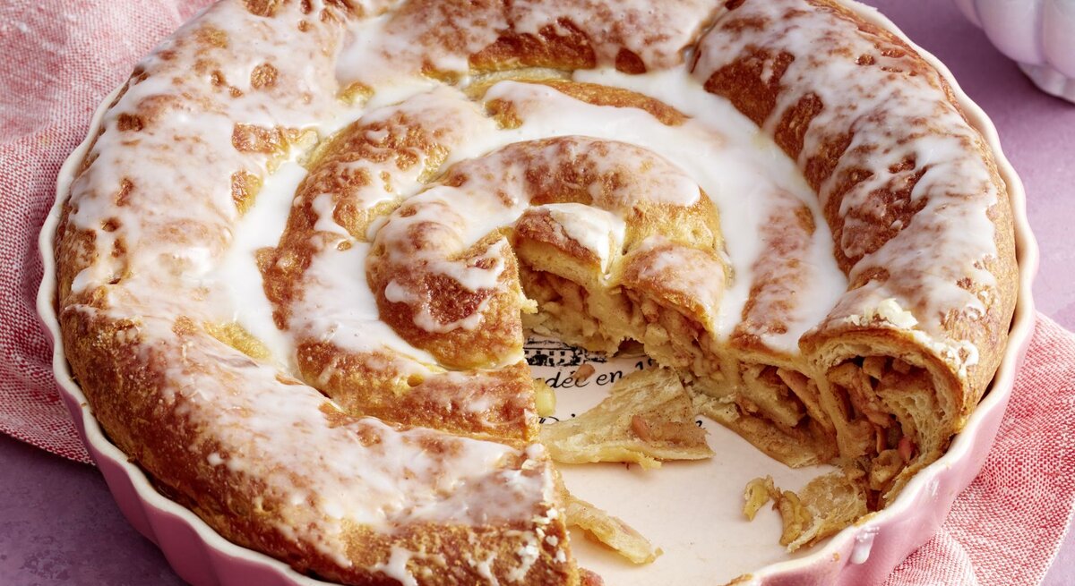Frisch gebackene Croissant-Apfel-Schnecke in einer rosa Backform, überzogen mit glänzendem Zuckerguss und gefüllt mit Zimt und Apfelstückchen, auf einem lila Hintergrund mit rosafarbenem Tuch.