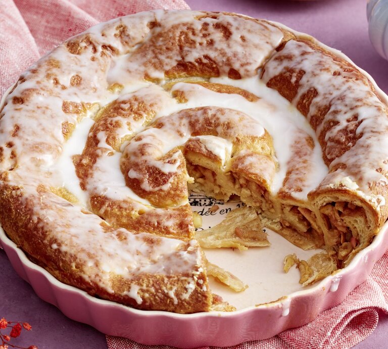 Frisch gebackene Croissant-Apfel-Schnecke in einer rosa Backform, überzogen mit glänzendem Zuckerguss und gefüllt mit Zimt und Apfelstückchen, auf einem lila Hintergrund mit rosafarbenem Tuch. | © EAT CLUB / Isabell Triemer