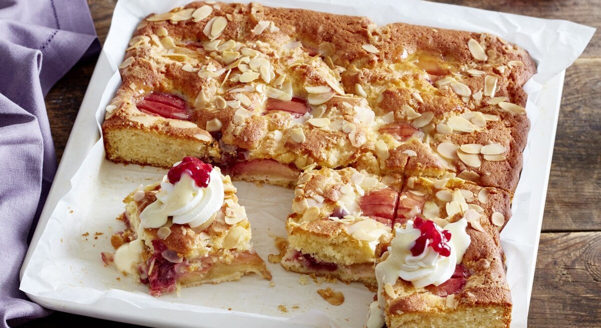 Saftiger Versunkener Apfel-Sahnekuchen auf einem Backblech, dekoriert mit Sahnetuffs, Preiselbeeren und gehobelten Mandeln, auf einem rustikalen Holztisch.
