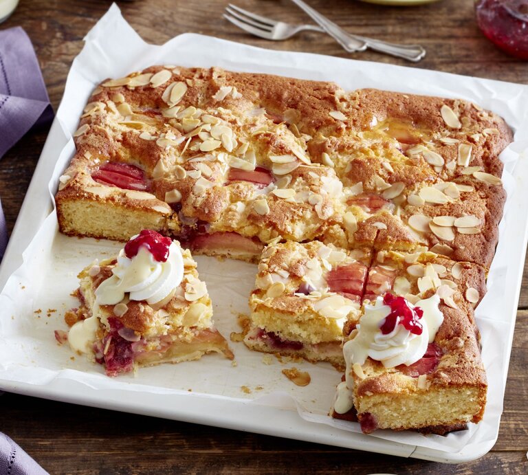 Saftiger Versunkener Apfel-Sahnekuchen auf einem Backblech, dekoriert mit Sahnetuffs, Preiselbeeren und gehobelten Mandeln, auf einem rustikalen Holztisch. | © EAT CLUB / Isabell Triemer