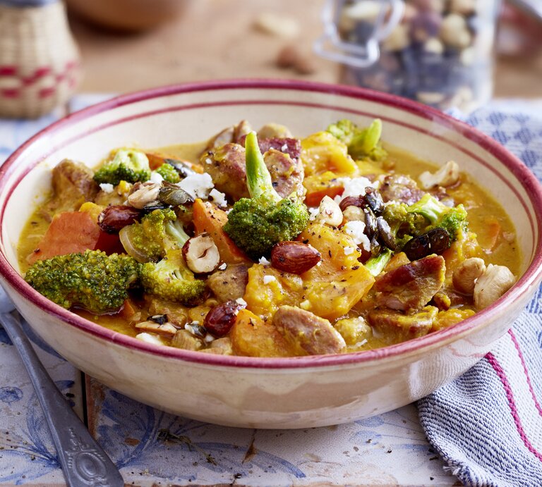 Puten-Gulasch mit Kürbis und Honignüssen in einer hellrosa Schüssel auf weiß-blauem Hintergrund. Rechts davon ein weiß-blau-kariertes Geschirrtuch und links von der Schüssel ein Löffel. | © EAT CLUB / Isabell Triemer