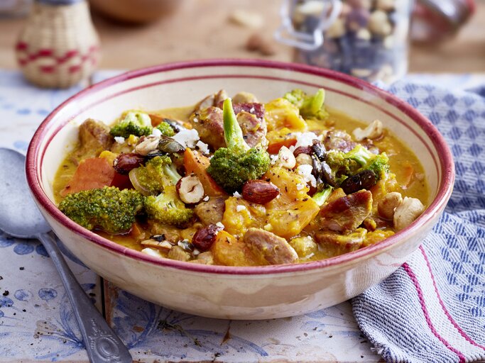 Puten-Gulasch mit Kürbis und Honignüssen in einer hellrosa Schüssel auf weiß-blauem Hintergrund. Rechts davon ein weiß-blau-kariertes Geschirrtuch und links von der Schüssel ein Löffel. | © EAT CLUB / Isabell Triemer
