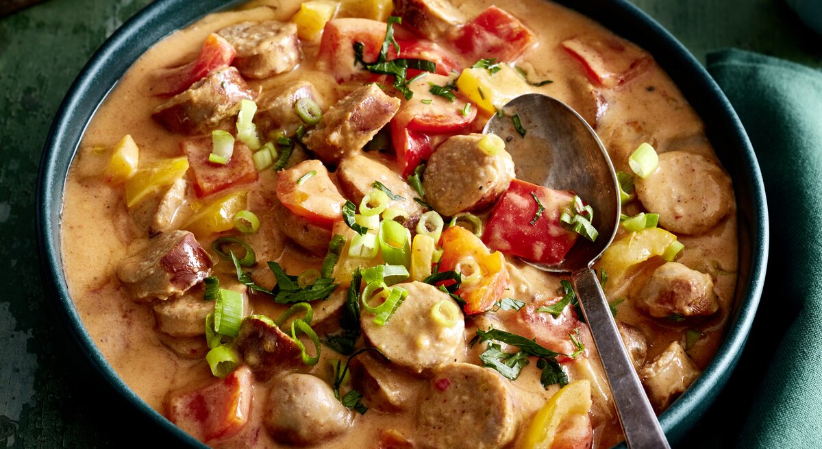 Wurstgulasch mit Paprika-Frischkäse-Soße in einer Schüssel auf dunkelgrünem Hintergrund. 