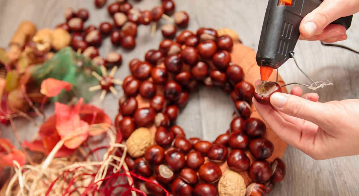 Person bastelt mit Kastanien, Walnüssen und rotem Bastband an einem herbstlichen Kastanienkranz auf grauem Holztisch.