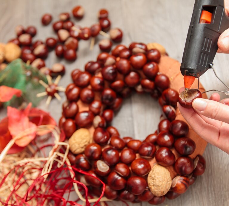 Person bastelt mit Kastanien, Walnüssen und rotem Bastband an einem herbstlichen Kastanienkranz auf grauem Holztisch. | © AdobeStock/rades