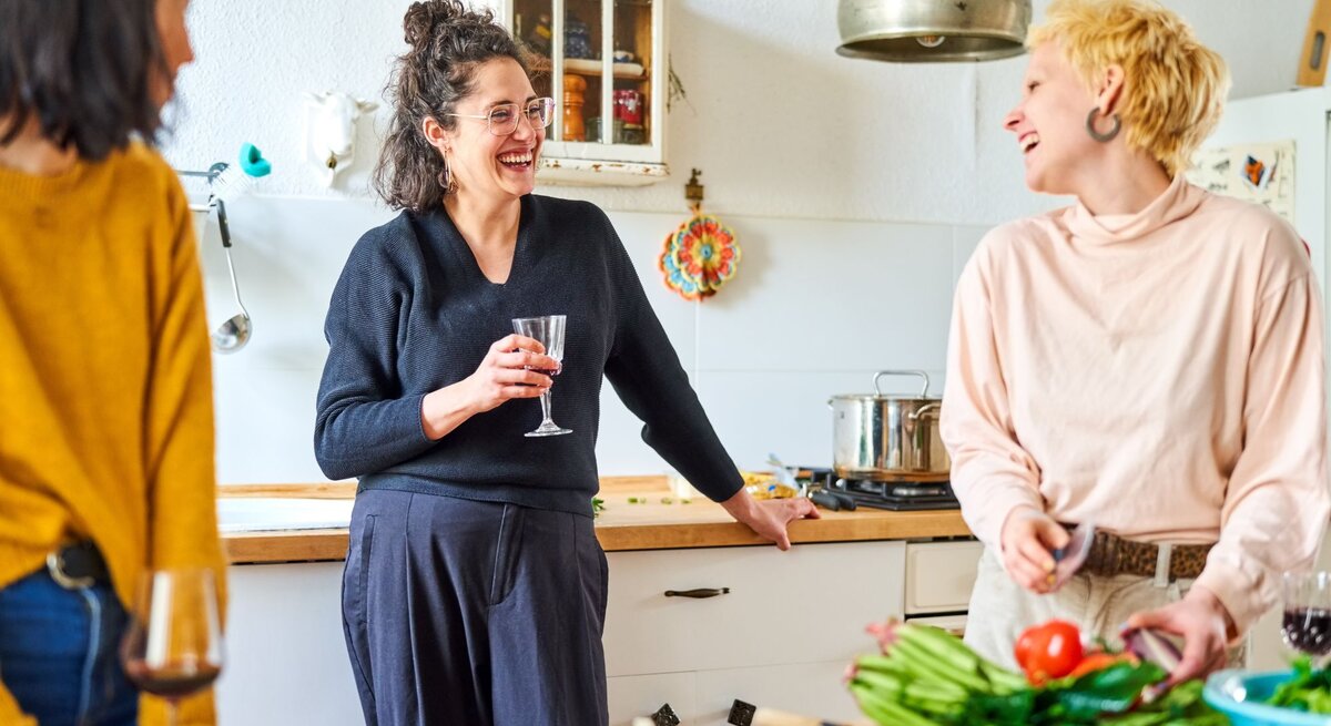 Drei Frauen stehen bei Vorbereitungen fürs Essen in der Küche. Sie unterhalten sich mit einem Glas Wein in der Hand und die Gemeinschaft macht ihnen Spaß.