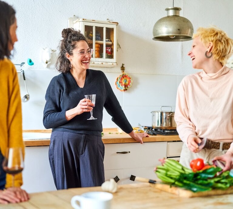 Drei Frauen stehen bei Vorbereitungen fürs Essen in der Küche. Sie unterhalten sich mit einem Glas Wein in der Hand und die Gemeinschaft macht ihnen Spaß. | © getty images/ Portra