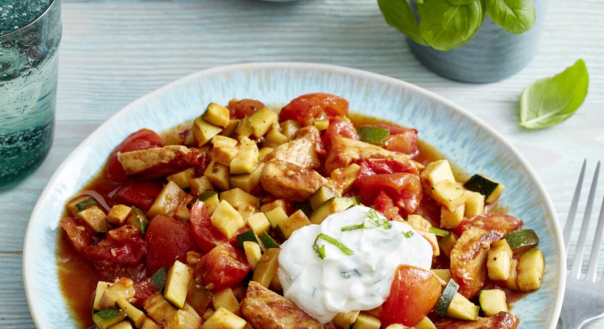 Steak-Streifen mit Tomaten-Zucchini-Ragout und frischem Kräuterquark angerichtet auf hellem Holz mit Basilikumdeko.