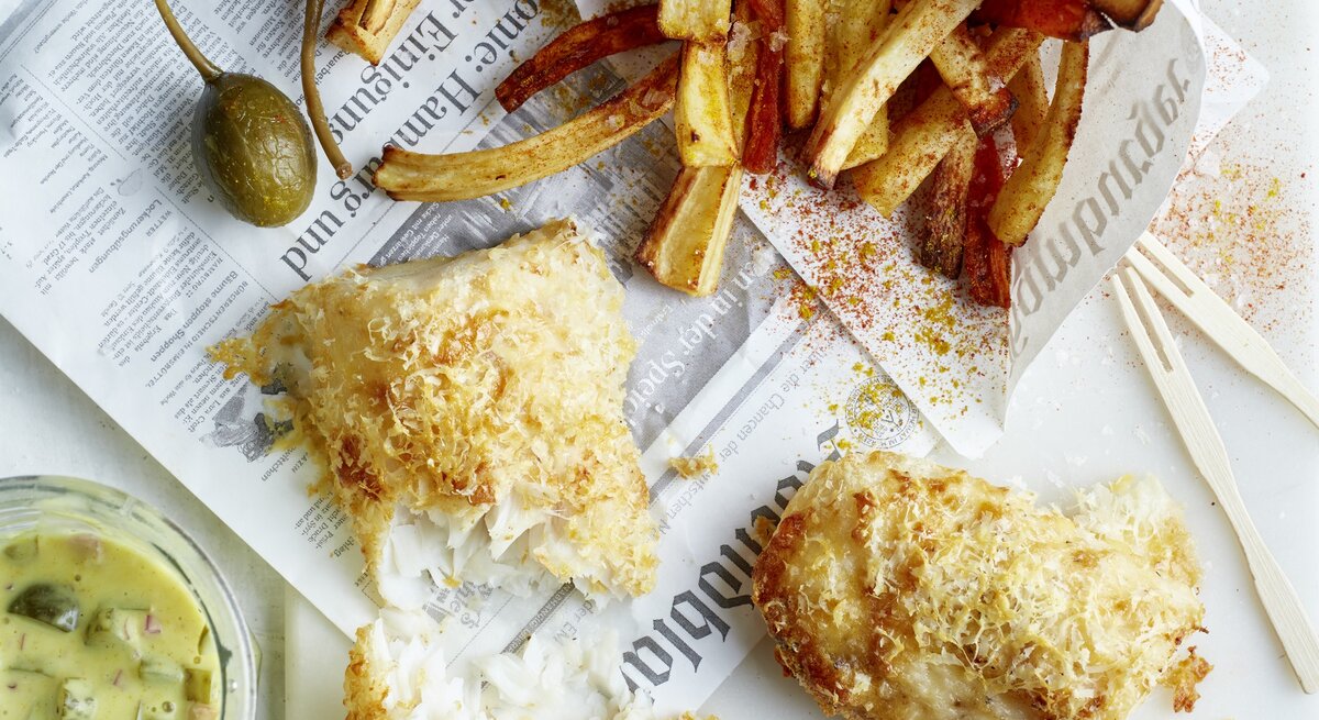 Rustikales Fish-and-Chips-Gericht mit Pommes in Papier, kross gebackenem Fisch, Zitronenspalten und Glas Remoulade.
