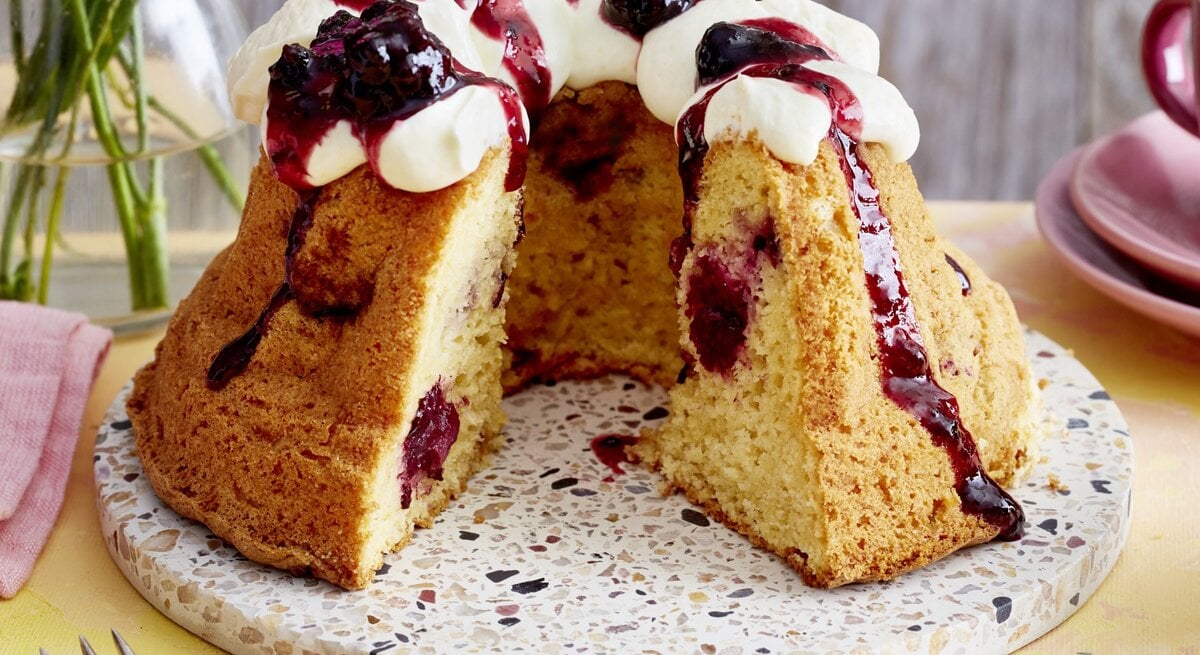 Brombeer-Apfel-Gugelhupf auf einem Tortenteller auf gelben Hintergrund. 