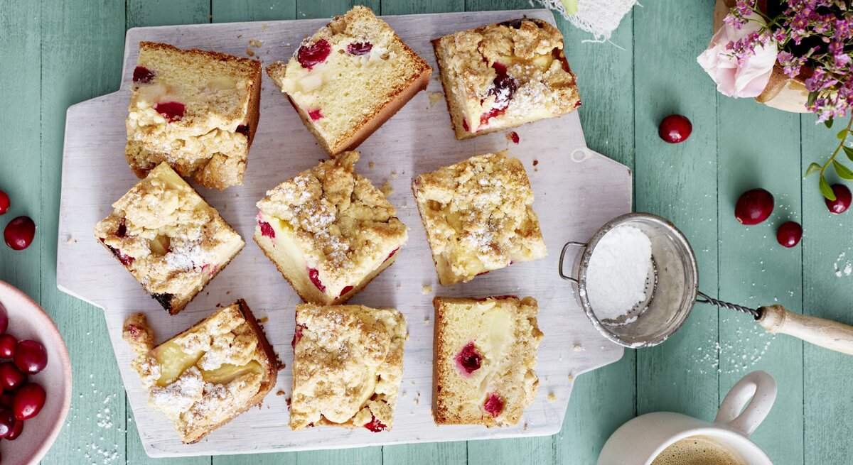Apfel-Cranberry-Streuselkuchen auf einem weißen Schneidebrett auf grünem Hintergrund.