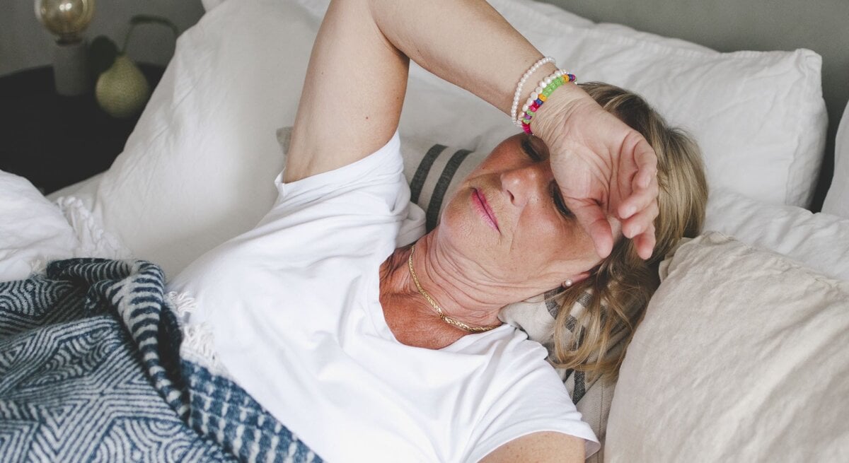 Zu sehen ist eine Frau mittleren Alters im Bett liegend, der sichtlich zu warm ist.