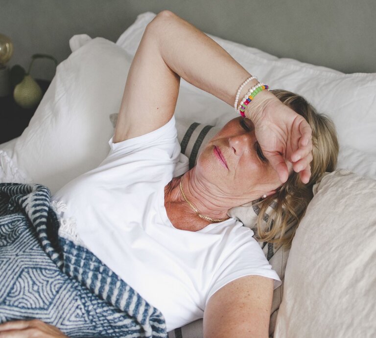 Zu sehen ist eine Frau mittleren Alters im Bett liegend, der sichtlich zu warm ist. | © GettyImages/Maskot