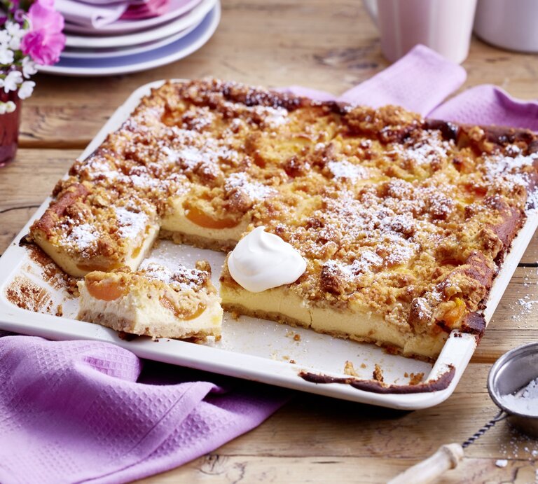 Aprikosen-Schmandkuchen auf einem weißen Backblech auf hölzernem Hintergrund. Links auf dem Foto ein Geschirrtuch und Blumen, rechts Puderzucker.  | © EAT CLUB/Isabell Triemer