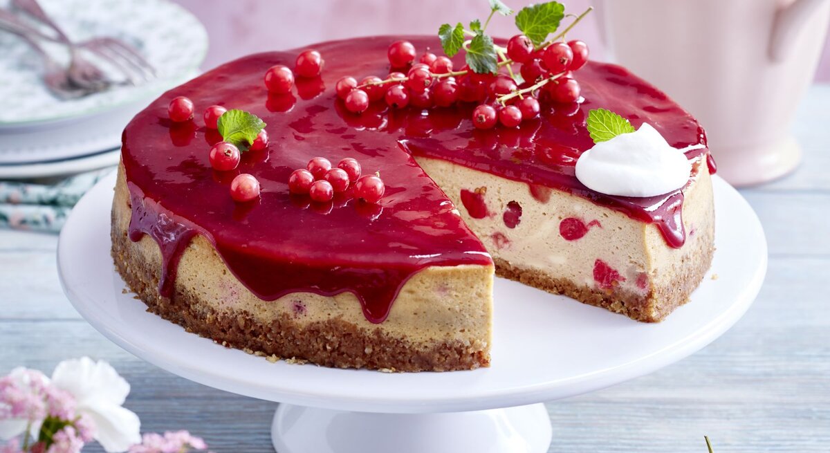  Johannisbeer-Cheesecake auf einem weißen Tortenteller vor hellem Hintergrund. Hinten auf dem Foto kleine Teller, vorne Blumen und Johannisbeeren.