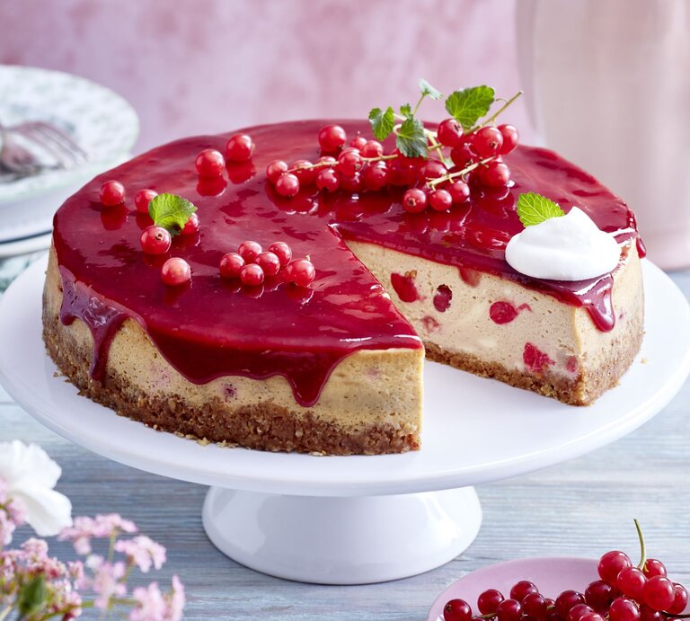  Johannisbeer-Cheesecake auf einem weißen Tortenteller vor hellem Hintergrund. Hinten auf dem Foto kleine Teller, vorne Blumen und Johannisbeeren. | © EAT CLUB/Isabell Triemer