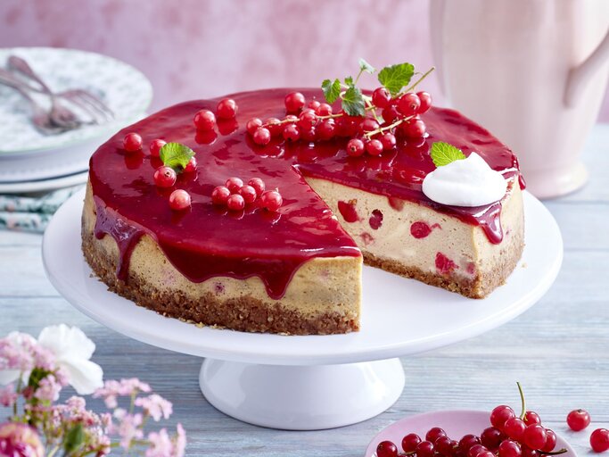  Johannisbeer-Cheesecake auf einem weißen Tortenteller vor hellem Hintergrund. Hinten auf dem Foto kleine Teller, vorne Blumen und Johannisbeeren. | © EAT CLUB/Isabell Triemer