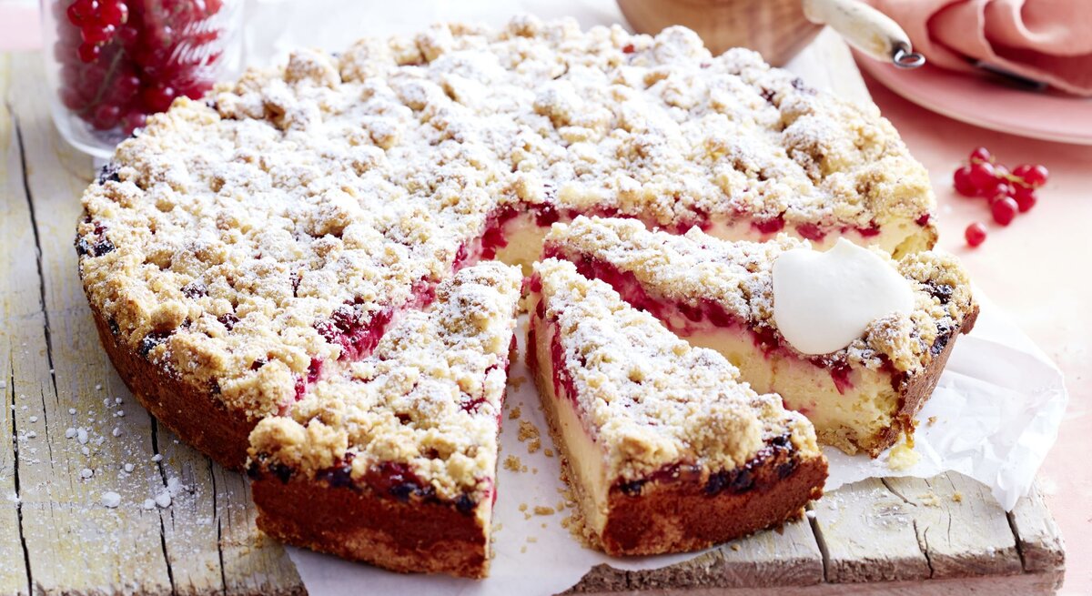 Johannisbeer-Käsekuchen mit Streuseln auf einem Holzbrett serviert, ein Stück angeschnitten, daneben ein Glas mit roten Johannisbeeren und Gänseblümchen.