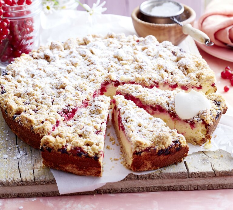 Johannisbeer-Käsekuchen mit Streuseln auf einem Holzbrett serviert, ein Stück angeschnitten, daneben ein Glas mit roten Johannisbeeren und Gänseblümchen. | © EAT CLUB/Isabell Triemer
